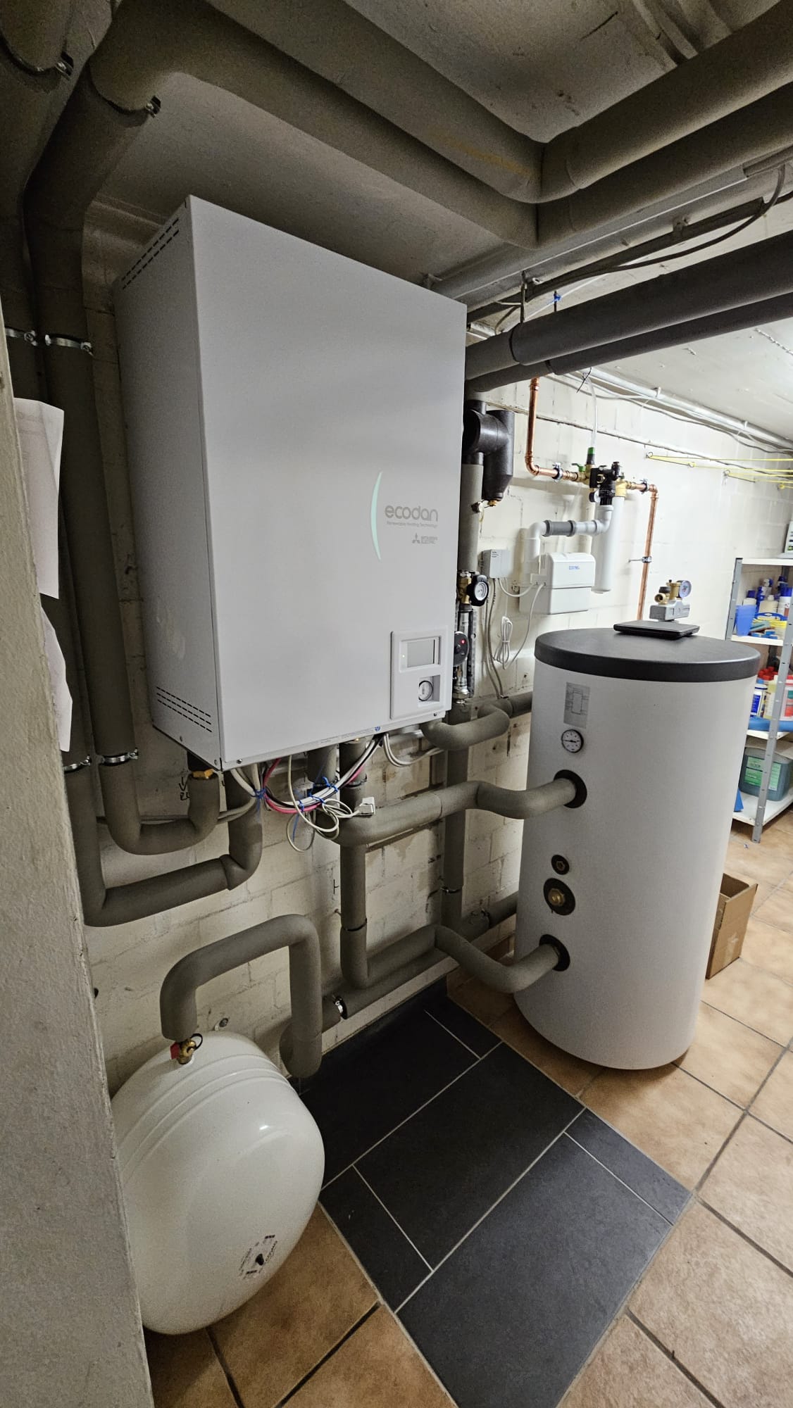 Moderne Heizungsanlage im Keller mit Wärmepumpen-Inneneinheit, Pufferspeicher, Rohrleitungen und Regeltechnik an der Wand.